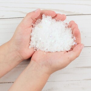May include: A pile of white artificial snow with iridescent, reflective flakes mixed throughout. The texture is granular and loose, resembling freshly fallen snow. The material appears lightweight and crystalline, catching the light with subtle rainbow-colored glints across the surface of the white particles.