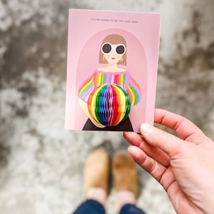 Op de afbeelding: Een roze kaart met de tekst "You're going to be the cool mom" en een illustratie van een vrouw met een zonnebril die een regenboog gestreept shirt draagt en een regenboog honingraatbal vasthoudt.
