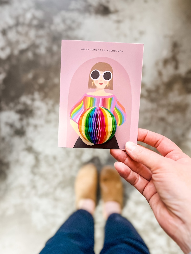 Op de afbeelding: Een roze kaart met de tekst "You're going to be the cool mom" en een illustratie van een vrouw met een zonnebril die een regenboog gestreept shirt draagt en een regenboog honingraatbal vasthoudt.