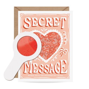 May include: A greeting card with the words "SECRET MESSAGE" in white and red lettering. A red heart with a lace design is in the center. A white magnifying glass is in front of the card. The text "REVEAL WITH THE DECODER SPY GLASS" is at the bottom.