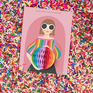 Op de afbeelding: Een roze kaart met een cartoonillustratie van een persoon met een zonnebril en een regenboog gestreept shirt dat een regenboog honingraatbal vasthoudt. De tekst op de kaart luidt "You're going to be the cool mom."