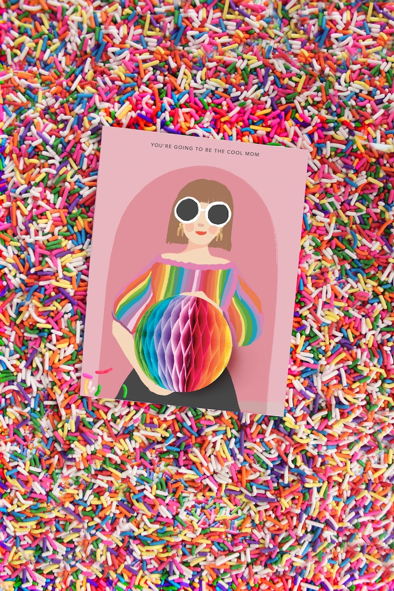 Op de afbeelding: Een roze kaart met een cartoonillustratie van een persoon met een zonnebril en een regenboog gestreept shirt dat een regenboog honingraatbal vasthoudt. De tekst op de kaart luidt "You're going to be the cool mom."