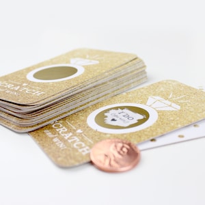 May include: A stack of gold glitter scratch off cards with the text "Scratch and Win" and a ring with a diamond. The cards have a gold glitter background with a white ring and diamond design. The top card has a circle to scratch off. A penny is used to scratch off the circle.