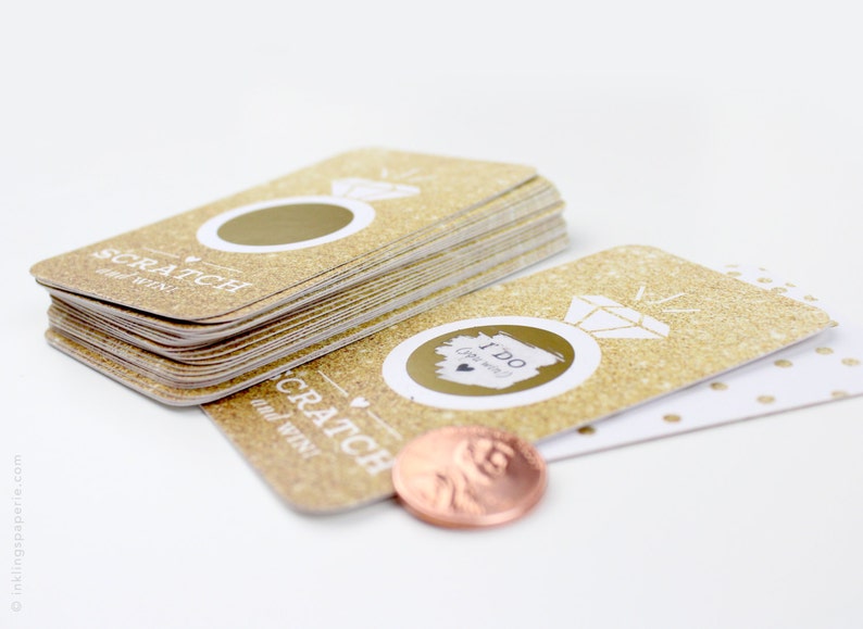 May include: A stack of gold glitter scratch off cards with the text "Scratch and Win" and a ring with a diamond. The cards have a gold glitter background with a white ring and diamond design. The top card has a circle to scratch off. A penny is used to scratch off the circle.
