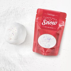 May include: A white, spherical snowball-like object with iridescent flecks next to a red, resealable pouch. The pouch features the text "inklings", "NORTH POLE Snow", and "JUST ADD WATER!" with a circular window showing white, glittery powder.
