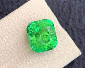 Puede incluir: Una vibrante gema verde esmeralda con talla cojín cuadrada. La gema se muestra sobre una tela beige claro, sobre un fondo gris oscuro. Las facetas de la gema reflejan la luz, mostrando su claridad y brillo.