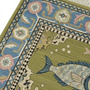 May include: Close-up of a decorative rug with a fish design. The rug features a green field with light blue fish and a border of blue, pink, and green floral patterns. The rug has a cream-colored fringe.