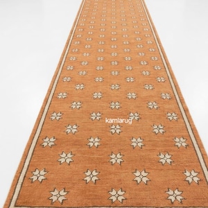 May include: An orange runner rug with a repeating floral pattern in off-white. The rug has a cream-colored border and a dark brown trim. The rug is made of a woven material and is suitable for hallways or entryways.