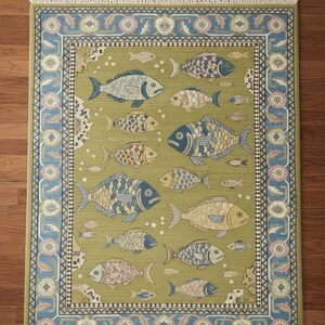 May include: A rectangular area rug with a fish pattern. The rug has a light green background with various fish in shades of blue, tan, and pink. A blue and pink border surrounds the fish design. The rug has a fringed edge.