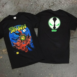 May include: Two black t-shirts featuring the comic book character Spawn. One shirt displays a colorful graphic of Spawn with the word "Spawn" in yellow and green. The other shirt has a green and white logo with the word "Spawn" in green.