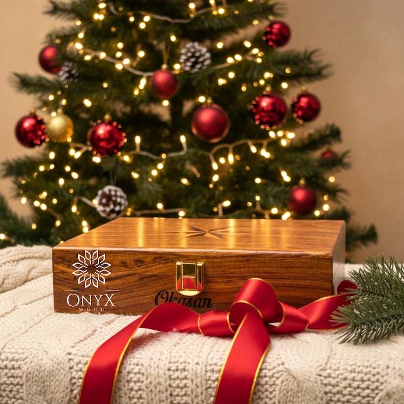 May include: A wooden box with the text "ONYX wood" and a gold clasp, resting on a white knitted blanket. A red ribbon is tied around the box. A Christmas tree with red ornaments and lights is in the background.