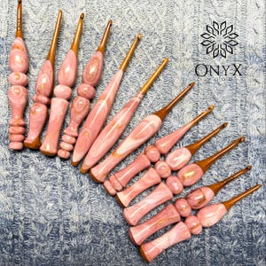Può includere: Una collezione di dieci uncinetti con manici in resina marmorizzata rosa liscia, caratterizzati da pagliuzze color oro e punte in legno. Gli uncinetti sono disposti in diagonale. Il logo Onyx Wood è visibile in alto a destra con un'icona floreale.