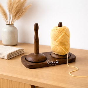 Può includere: Un porta filo in legno marrone scuro con due montanti verticali. Un montante tiene una palla di filo giallo. La base rettangolare ha il testo "ONYX WOOD" in bianco. Un vaso con fiori secchi e un panno piegato sono sullo sfondo.