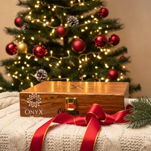 May include: A rectangular wooden box with a polished brown grain finish, featuring a gold-colored metal latch, a star-shaped inlay on the lid, and the text ONYX WOOD and Okasan printed on the front, accented by a red ribbon and a pine branch.