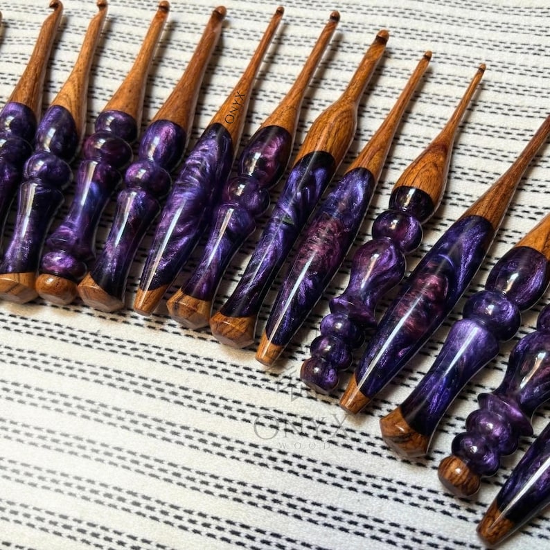 May include: A collection of crochet hooks featuring turned wooden handles combined with marbled purple resin. Each hook displays a unique grain pattern in the wood and swirling violet tones within the resin, with some handles showing the word Onyx.