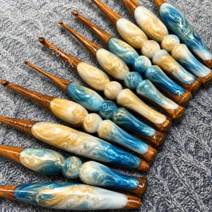 May include: A collection of crochet hooks featuring wooden shafts and handles crafted from a blend of blue, white, and gold swirled resin. Each handle displays a unique contoured shape with rounded sections. The word ONYX is printed on the hooks.