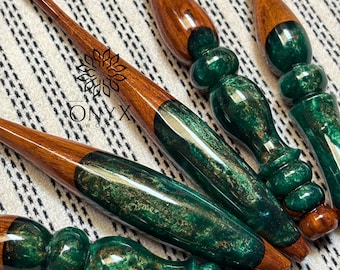 Könnte beinhalten: Eine Sammlung handgefertigter Häkelnadeln mit Griffen aus poliertem braunem Holz und Abschnitten aus marmoriertem smaragdgrünem Harz mit Goldpartikeln. Die Nadeln zeigen ein Logo mit dem Text ONYX WOOD. Das Design kombiniert glatte Holzspitzen mit ergonomischen, strukturierten Harzgriffen.