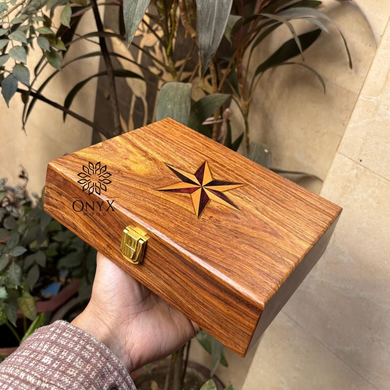 May include: A rectangular wooden box with a compass rose inlay and a gold-colored clasp. The box is made of a warm-toned wood and features the text "ONYX WOOD". The compass rose is a starburst design with red and gold accents.