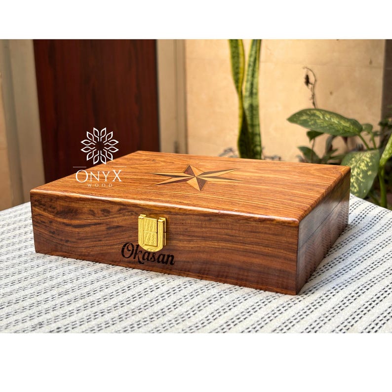 May include: A rectangular wooden box with a compass rose design on the lid. The box is made of dark brown wood and has a gold-colored clasp. The text "ONYX WOOD" and "Okasan" are visible on the box.