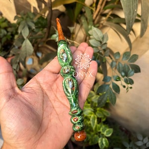 Puede incluir: Un ganchillo hecho a mano con un mango de madera y un gancho de metal. El mango está pintado en tonos verdes y marrones con un acabado brillante. El texto "ONYX WOOD" es visible en el gancho.