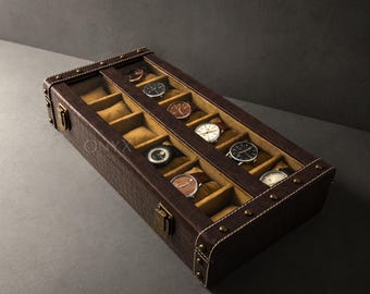 May include: A brown leather watch case with a clear glass top, showcasing multiple wristwatches. The case has a textured exterior, gold-colored hardware, and a cream-colored interior. The watches are displayed on individual cushions.