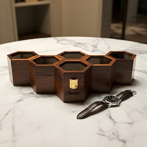 Puede incluir: Un organizador hexagonal de madera oscura con seis compartimentos, un cierre dorado y la palabra "Sopran" grabada en la parte delantera. Un reloj con esfera negra y correa de cuero marrón descansa sobre la superficie de mármol.