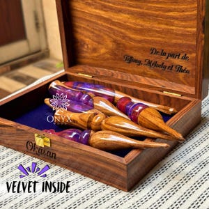 May include: A wooden box with a velvet interior holds several wooden crochet hooks with purple and red accents. The box is engraved with the words "Okasan" and "VELVET INSIDE". The lid is inscribed with "De la part de Tiffany, Melody et Theo."