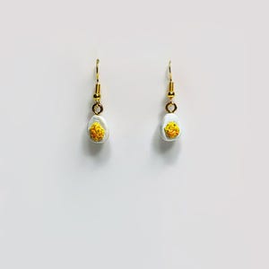 May include: A pair of handmade earrings featuring a halved egg design. The earrings have gold-colored hooks and small, detailed egg halves with white and yellow coloring. The earrings are set against a plain white background.