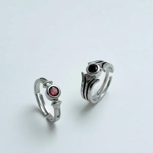 May include: Two silver rings with adjustable bands. One ring features a round, red gemstone, while the other has a black gemstone and black stripes. The rings are displayed on a white background.