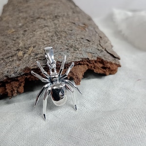 Puede incluir: Colgante de araña plateada pulida con detalles en patas y cuerpo. El colgante refleja la luz. Diseño realista, ideal para collar o charm. Se muestra sobre una superficie texturizada.