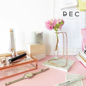 May include: A pink flower arrangement in a glass vase on a marble stand with a copper frame. The vase is on a pink surface with a calendar, books, and other decorative items.