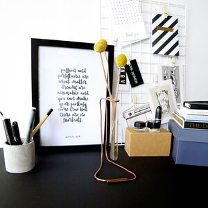 May include: A black and white framed print with the text "passion and persistence are what matter. dreams are achievable and you can make your fantasy come true but there are no shortcuts."  The print is on a black desk with a copper test tube holder with two yellow flowers.