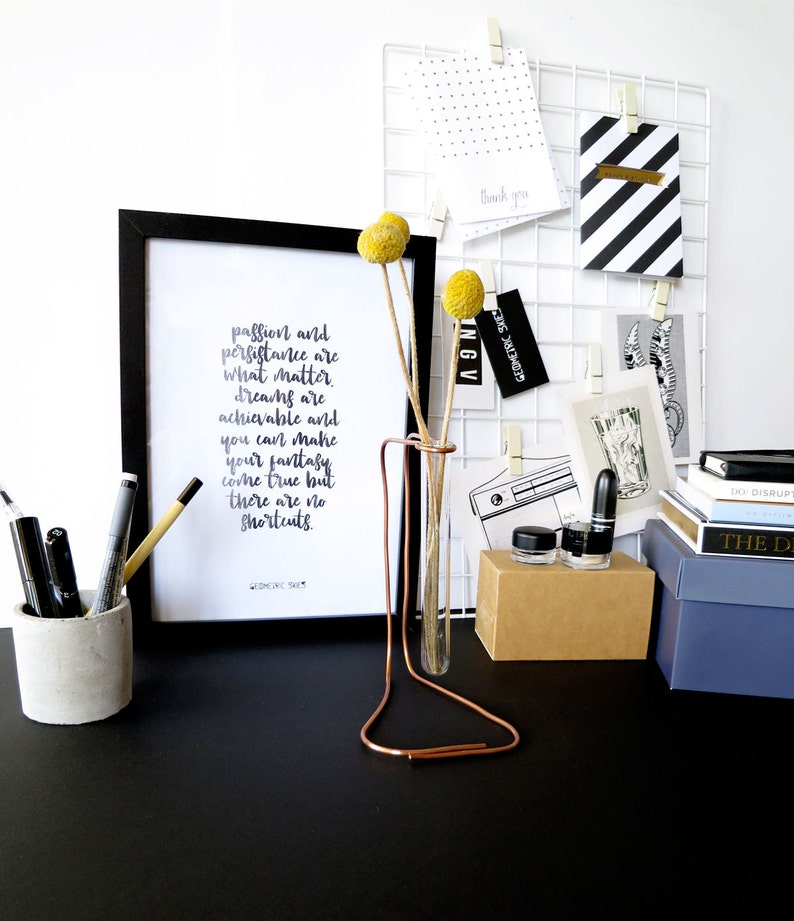 May include: A black and white framed print with the text "passion and persistence are what matter. dreams are achievable and you can make your fantasy come true but there are no shortcuts."  The print is on a black desk with a copper test tube holder with two yellow flowers.