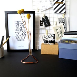 May include: A black framed print with a white background and black text that reads "passion and persistence is what matters. Dreams are achievable and you can make your fantasy come true but there are no shortcuts. -  unknown". A copper wire stand holds a glass vase with dried yellow flowers.