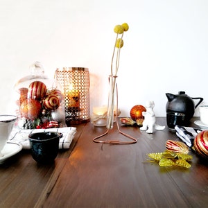May include: A table setting with a white ceramic teacup and saucer, a black ceramic cup, a glass cloche with red and gold Christmas ornaments, a gold metal candle holder, a glass vase with dried yellow flowers, a white ceramic figurine, a black teapot, a white ceramic saucer with a blue rim, and a red and gold Christmas ornament.