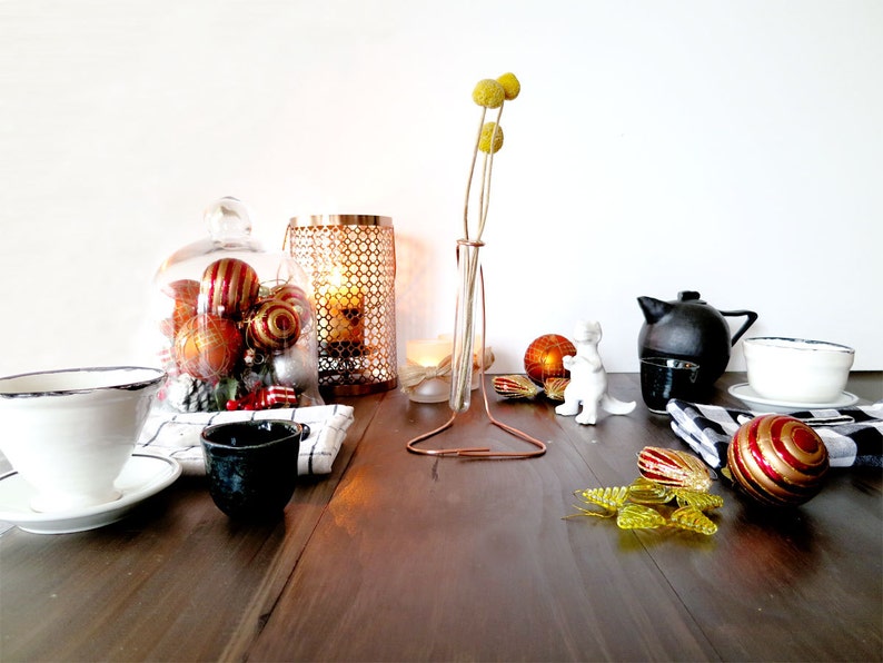 May include: A table setting with a white ceramic teacup and saucer, a black ceramic cup, a glass cloche with red and gold Christmas ornaments, a gold metal candle holder, a glass vase with dried yellow flowers, a white ceramic figurine, a black teapot, a white ceramic saucer with a blue rim, and a red and gold Christmas ornament.