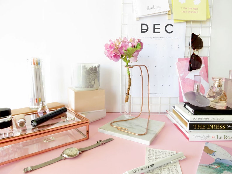 May include: A pink flower arrangement in a glass vase on a marble stand with a copper frame. The vase is on a pink surface with a calendar, books, and other decorative items.