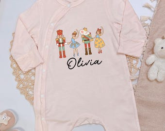 May include: A light pink baby romper with long sleeves and snap closures. The romper features a graphic of a nutcracker, a ballerina, a mouse king, and another ballerina. The name "Olivia" is written below the graphic.