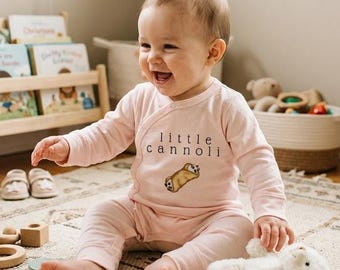Puede incluir: Un conjunto de bebé de color rosa claro con mangas largas, que presenta un gráfico de un cannoli y el texto "little cannoli" impreso en negro en el pecho. La prenda tiene un cierre cruzado con botones a presión y pantalones a juego.