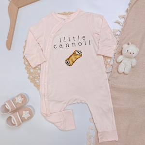 May include: A light pink baby romper with long sleeves and legs. The romper features the text "little cannoli" and an illustration of a cannoli pastry. A pair of pink baby shoes and a small white teddy bear are also visible.