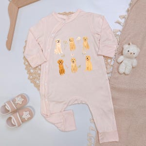 May include: A light pink long-sleeve baby romper featuring a graphic print of six golden retriever dogs and small white flowers. The garment has a side-button closure with white buttons running diagonally from the neckline to the waist.