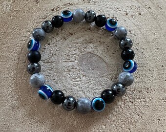 May include: A beaded bracelet featuring a mix of black, gray, and blue beads. The bracelet includes beads with an eye design, along with solid black and gray stone beads. The bracelet is displayed on a wooden surface.