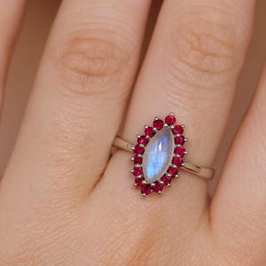 Könnte beinhalten: Ein silberner Ring mit einem ovalen, schillernden Mondstein in der Mitte, umgeben von kleinen, runden, roten Edelsteinen. Der Ring sitzt auf einem schmalen Silberband und präsentiert ein klassisches und elegantes Design.