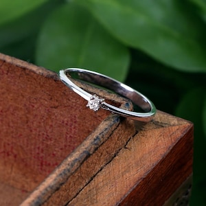 May include: A delicate silver ring with a small, sparkling gemstone. The ring is set on a wooden surface, with a blurred green background. The gemstone is the focal point, catching the light and adding a touch of elegance to the simple design.