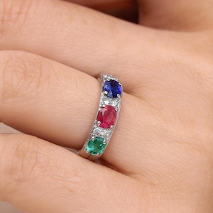 Puede incluir: Un anillo de plata con tres piedras preciosas ovales: azul, rojo y verde. Pequeñas piedras transparentes se colocan entre las gemas más grandes. El anillo se muestra en un dedo.