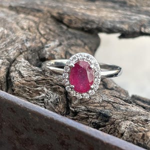 Peut inclure: Une bague en argent ornée d'une pierre précieuse ovale rouge foncé, entourée d'un halo de petites pierres claires. La bague est posée sur une surface en bois brun texturé, mettant en valeur l'éclat et le design du bijou.