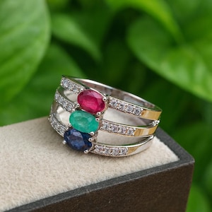 Puede incluir: Un anillo plateado con tres piedras preciosas ovaladas en rojo, verde y azul. El anillo tiene tres bandas, cada una con pequeñas piedras transparentes. El anillo se presenta sobre un fondo neutro con un fondo verde borroso.