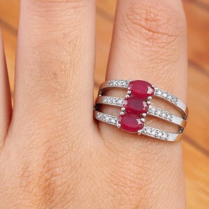 May include: A silver ring featuring three oval-cut, deep red gemstones, accented with small, clear stones. The ring has a multi-band design, with the gemstones centered on the top band.