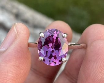 Può includere: Un anello d'argento con una grande pietra preziosa ovale nei toni del viola e del rosa. La gemma è tenuta da quattro griffe. L'anello è tenuto tra due dita.