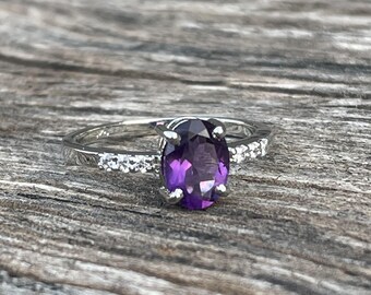 May include: A silver ring with a large, oval-cut, deep purple gemstone as the focal point. The band is adorned with smaller, clear stones on either side of the main stone, set against a wooden background.